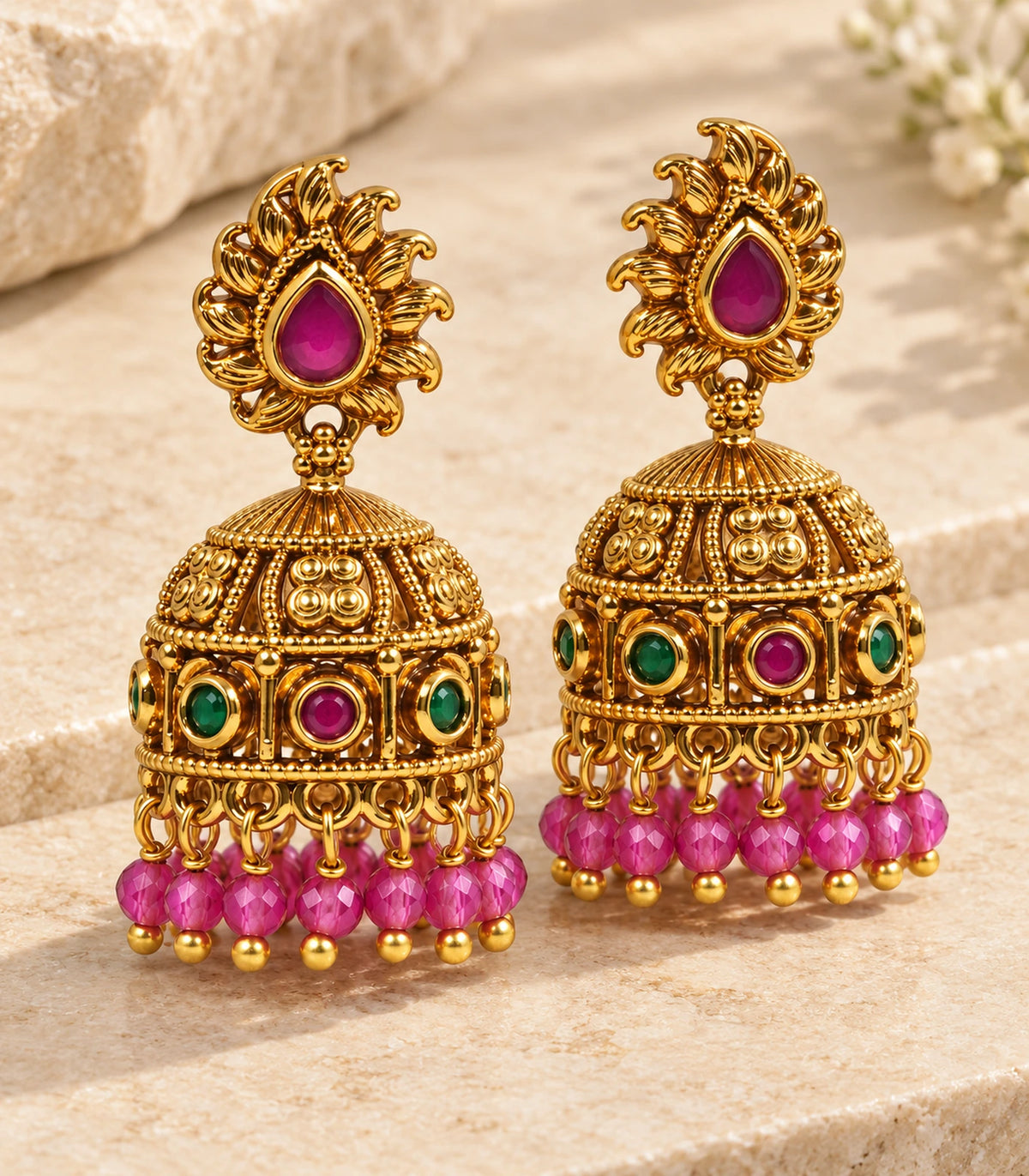 Pair of antique gold-tone magenta and green flame jhumka earrings on a neutral textured surface