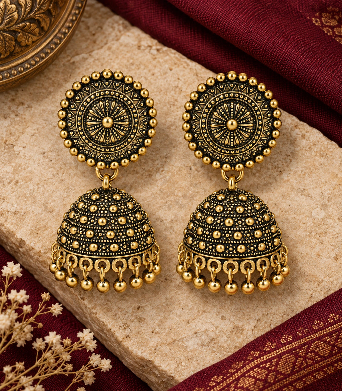 JIMIKI Antique Gold Sunburst Medallion Jhumka Earrings - Matching pair on pale stone beside maroon fabric #color_antique-gold