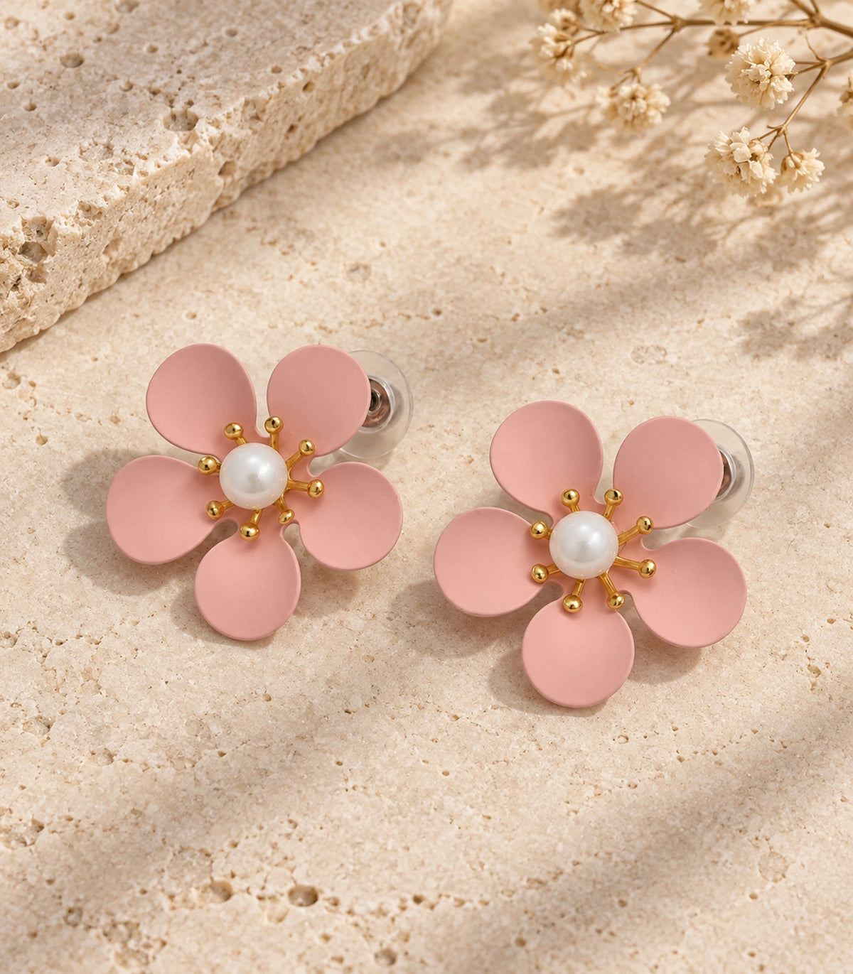 JIMIKI blush pink five-petal flower stud earrings with pearl-effect centre, matching flower stud pair in an editorial flat lay #color_blush-pink