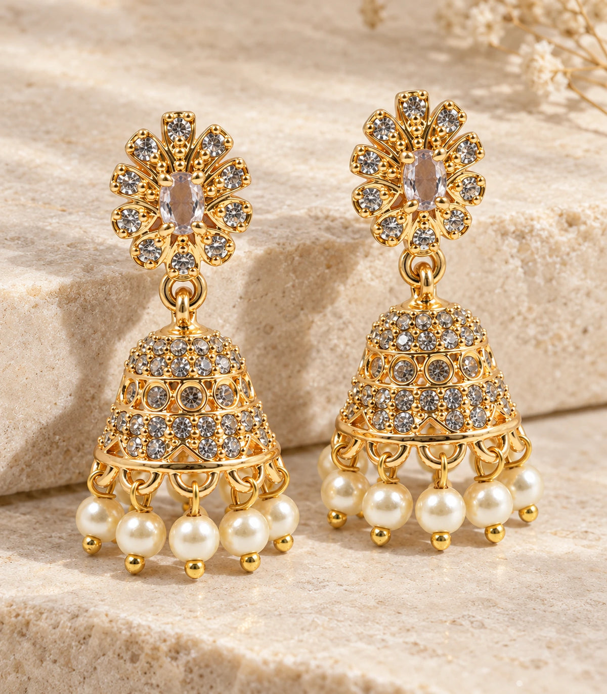 Complete matching pair of gold-tone champagne floral jhumka earrings styled on a beige stone surface