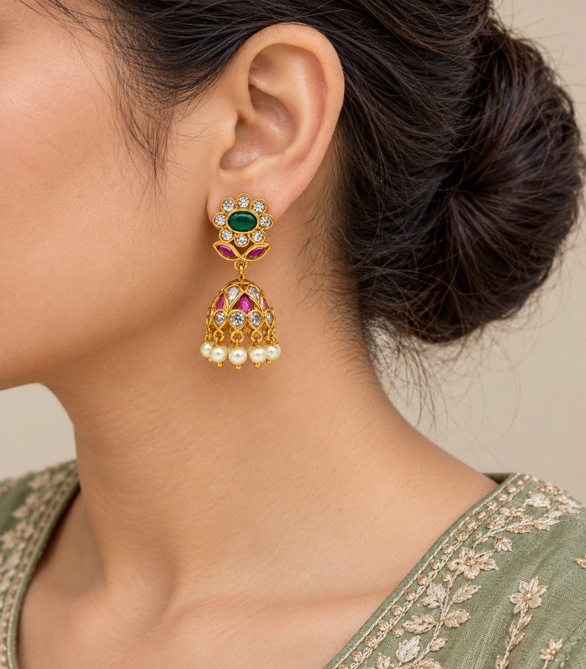 Closeup of one worn gold-tone floral jhumka with emerald-green oval centre, magenta bell accents and pearl fringe