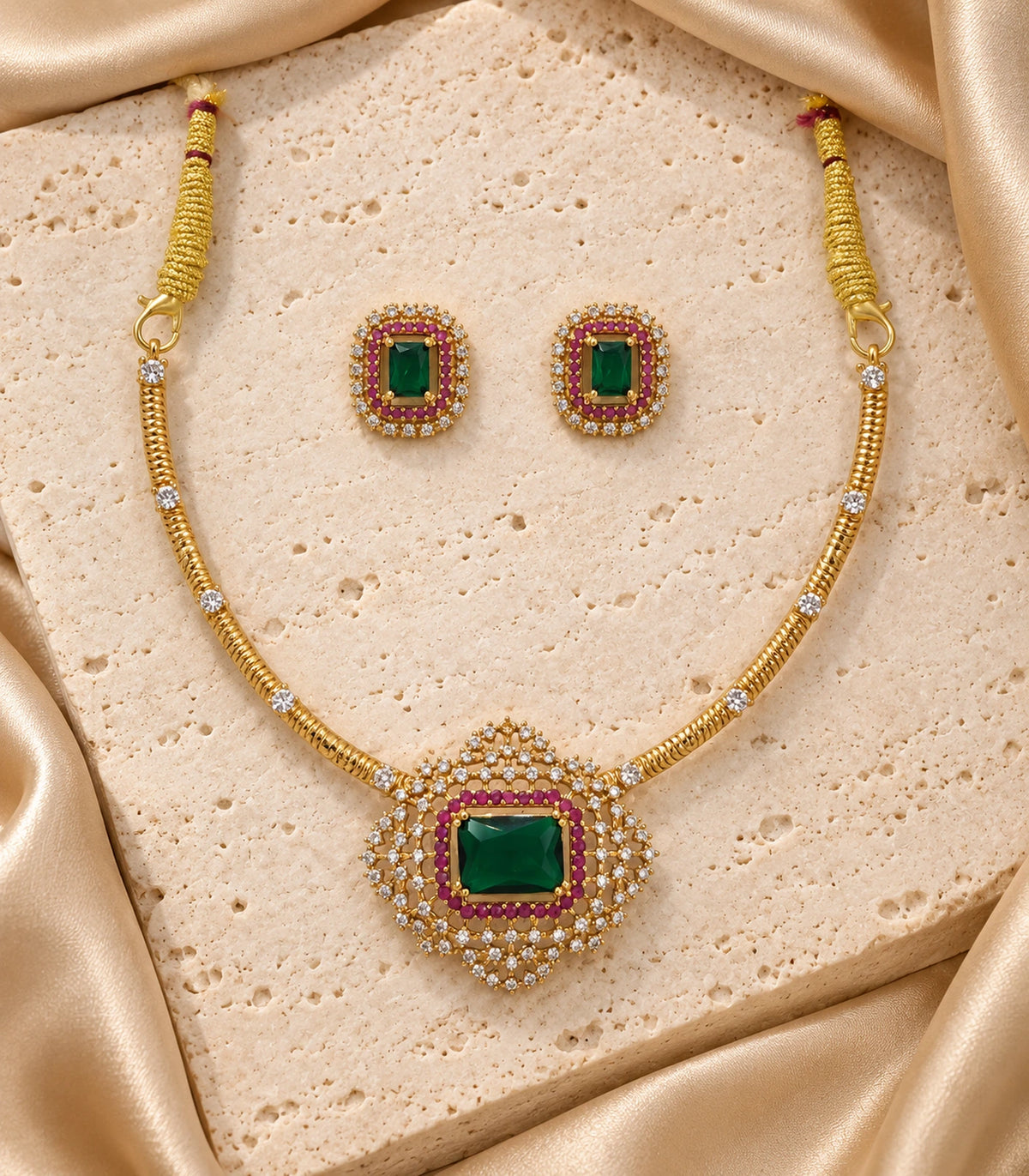Gold-colour necklace and emerald-green matching stud pair arranged on stone with cream fabric
