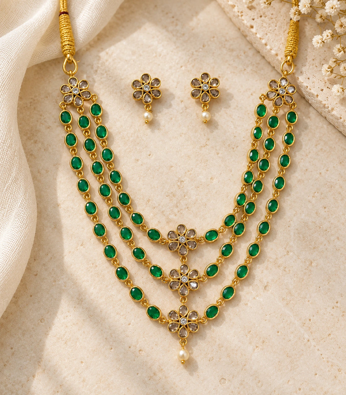 Gold-tone emerald-green triple-strand necklace and matching pair of floral white-drop earrings arranged on a cream surface