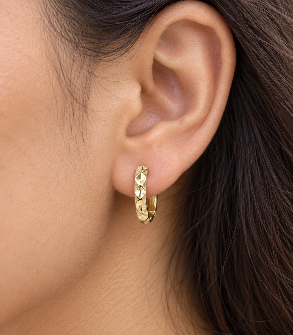 JIMIKI gold faceted diamond pattern huggie earrings, worn closeup of the front design #color_gold-tone