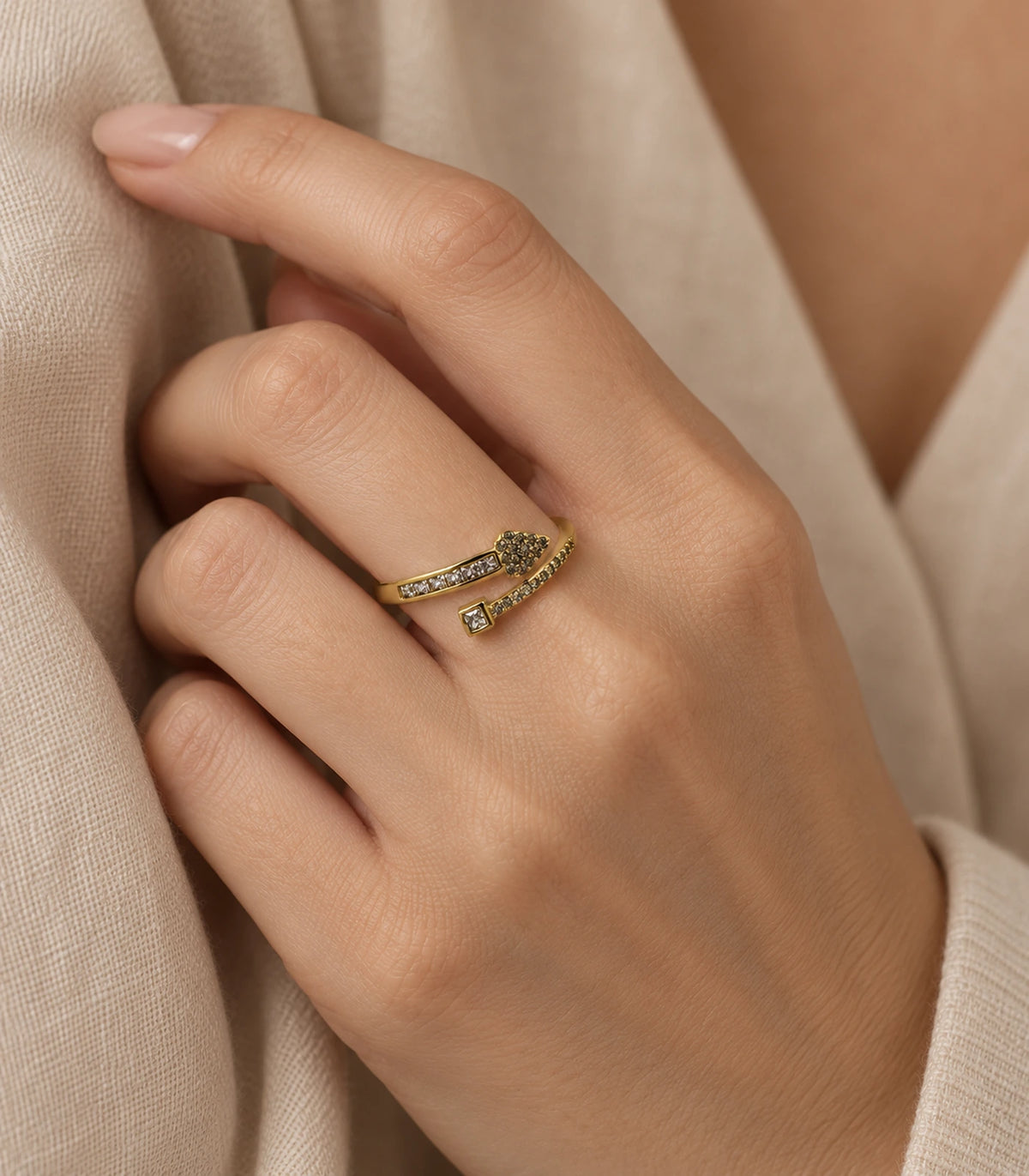 Gold heart and square wrap ring worn in a curled-hand pose with cream clothing