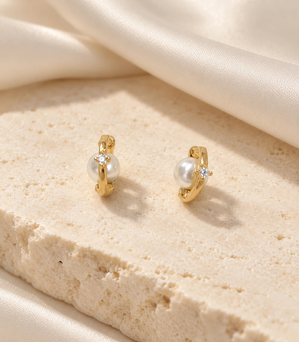 Matching pair of gold-tone pearl-style and clear-stone orbit huggie earrings on a stone surface with fabric