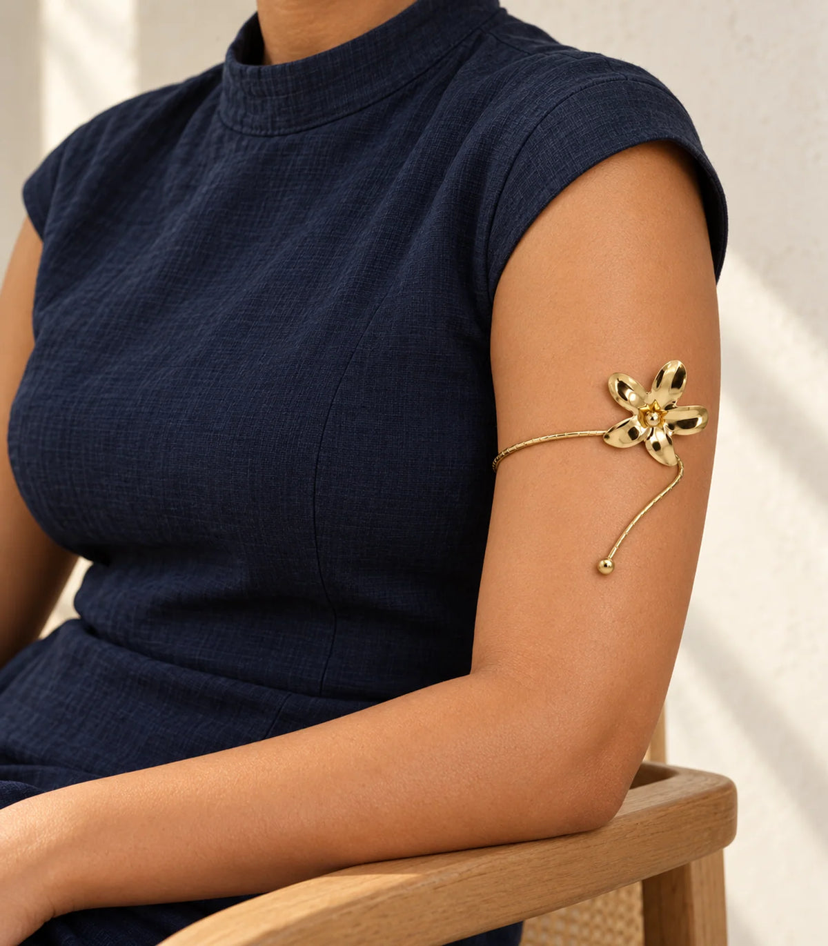 Gold-tone five-petal flower and ball-tip arm cuff on the upper arm in a seated pose with navy high-neck clothing