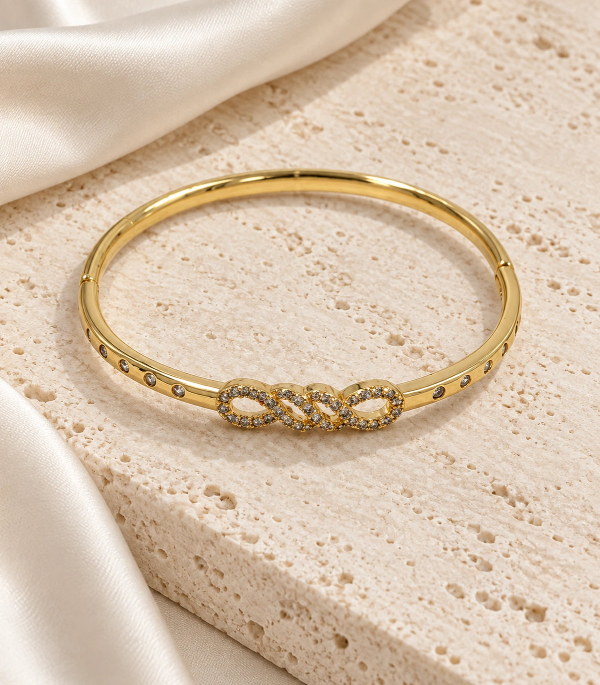 JIMIKI Gold-Tone Infinity Knot Bezel Stone Bangle — bangle in a styled flat lay #color_gold-tone / clear