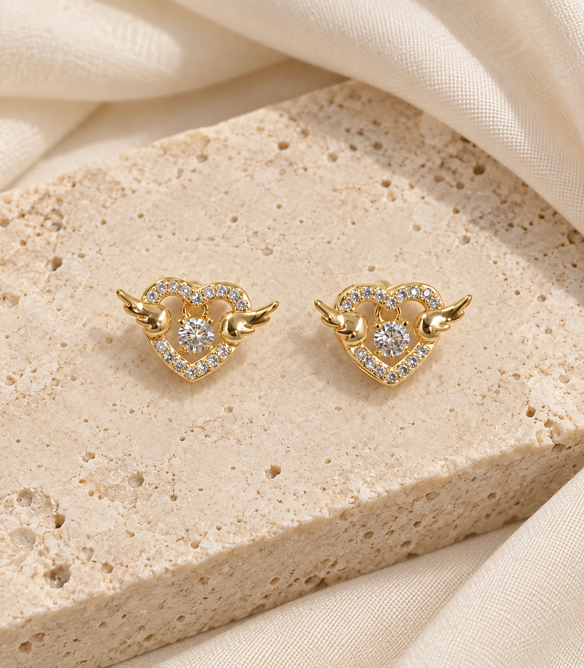 JIMIKI Winged Heart Crystal Stud Earrings in Gold Tone — editorial flat lay #color_gold-tone