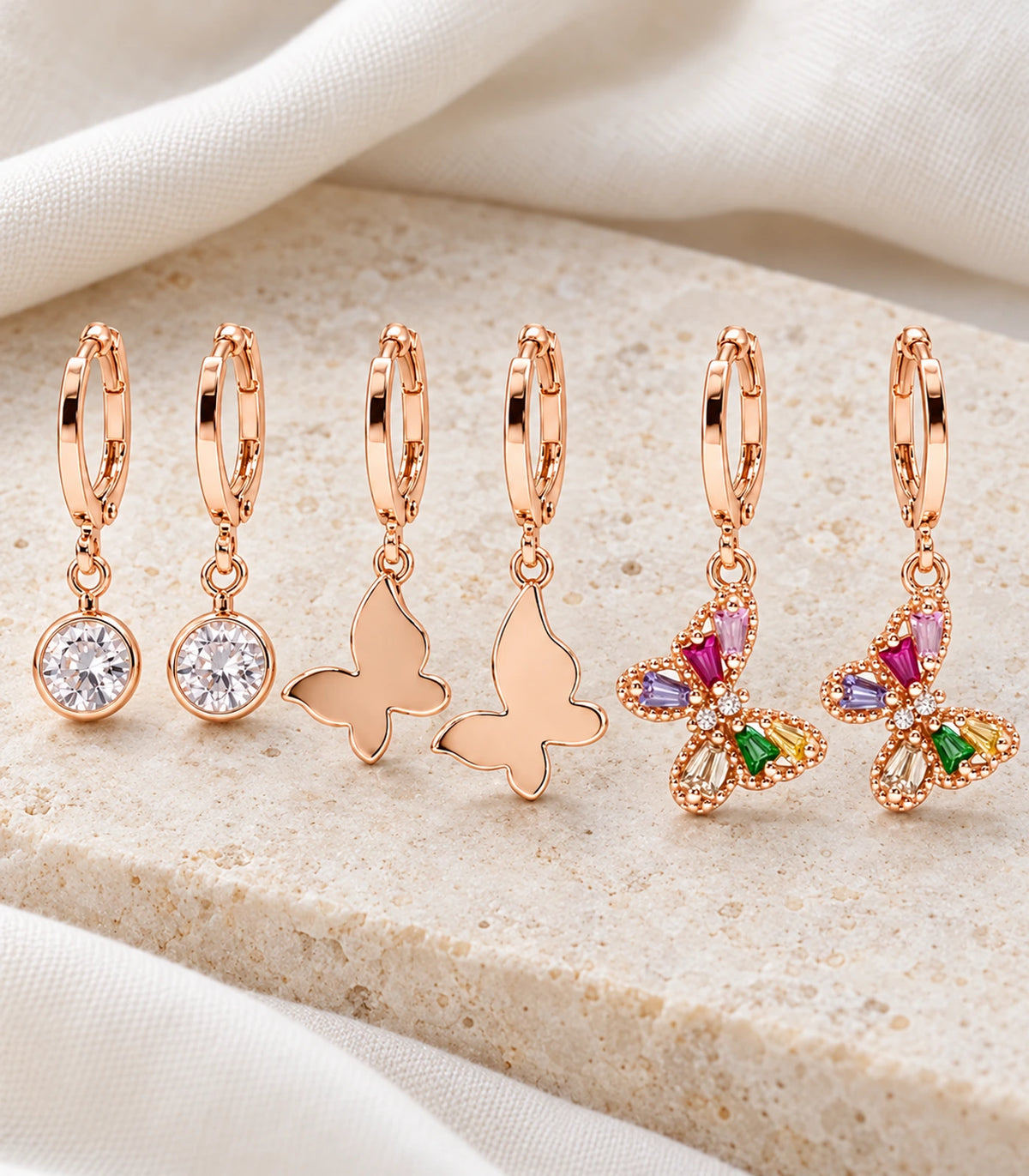 Complete three-pair rose gold tone crystal and butterfly hoop set on ivory fabric; six earrings