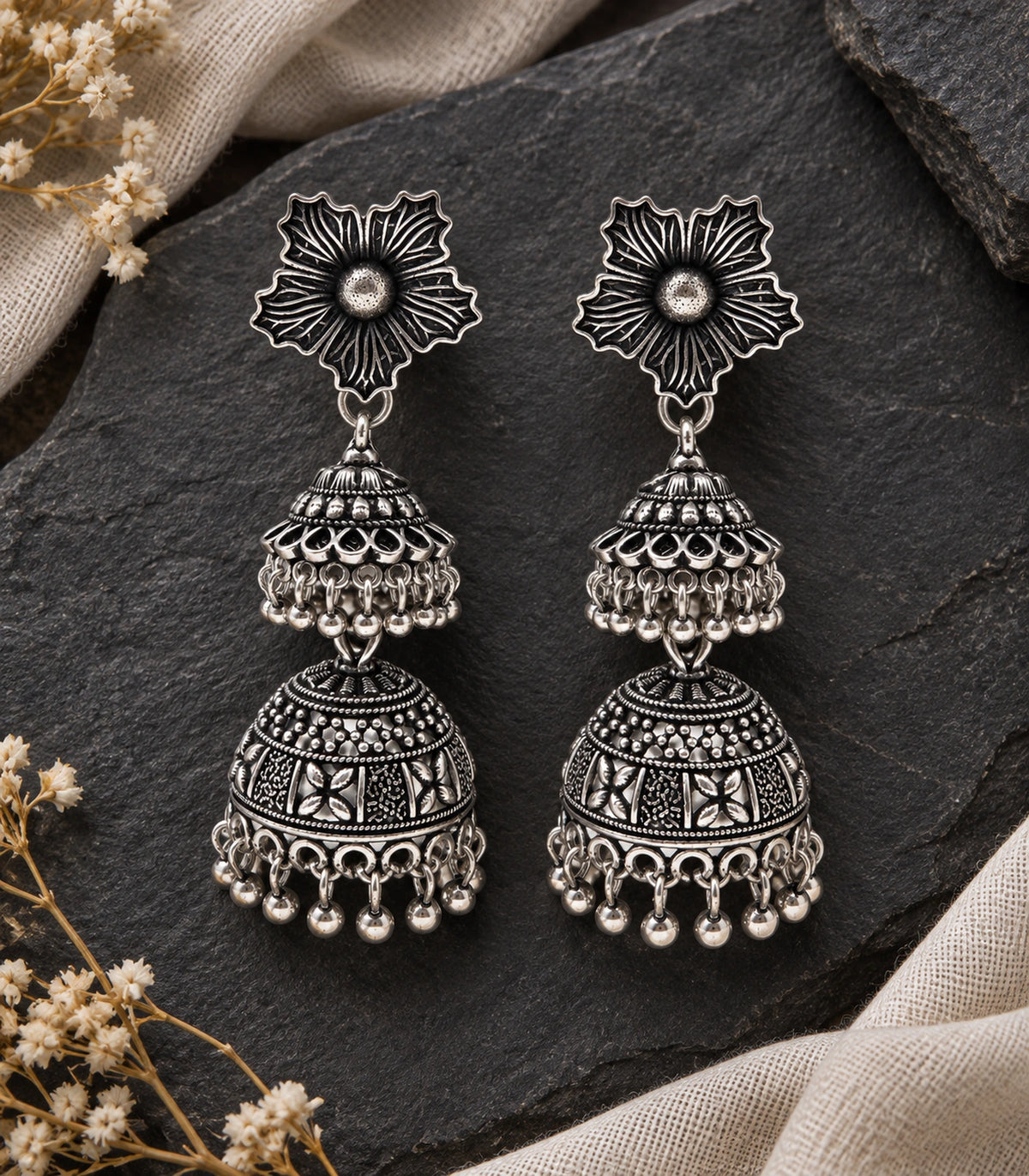 JIMIKI Oxidized Silver Hibiscus Double Jhumka Earrings - pair styled on dark stone with linen #color_oxidized-silver