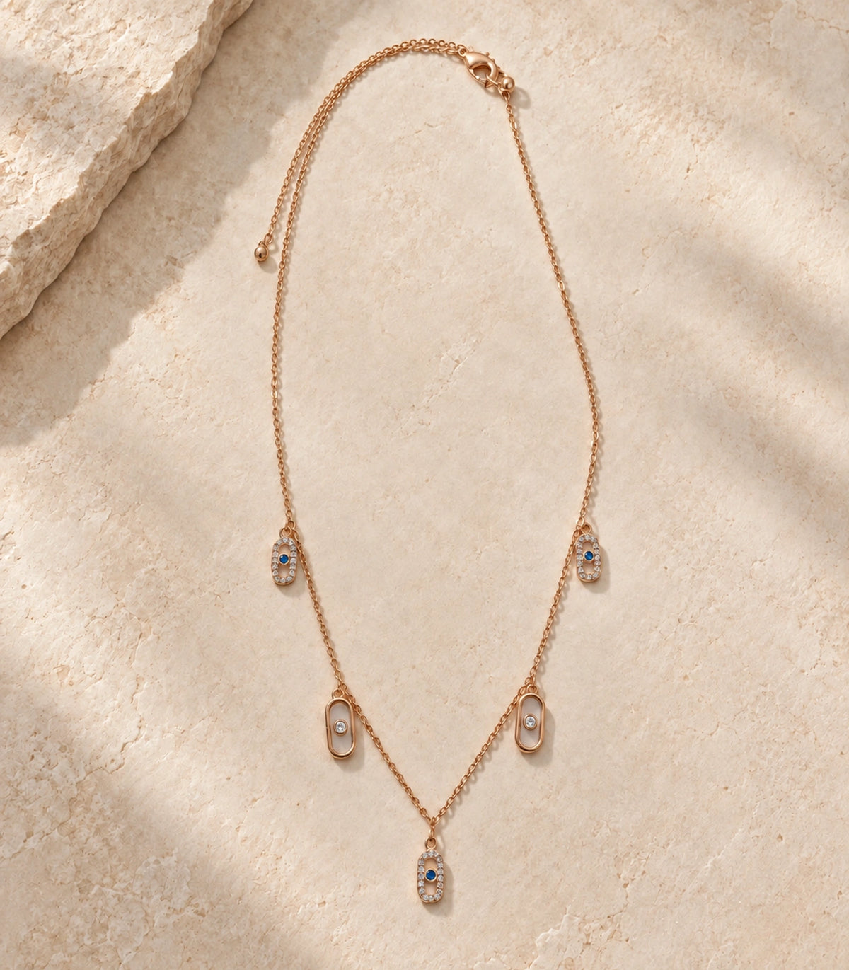 JIMIKI Rose Gold Blue Clear Oval Station Necklace - complete five-oval necklace in a styled flat lay #color_rose-gold
