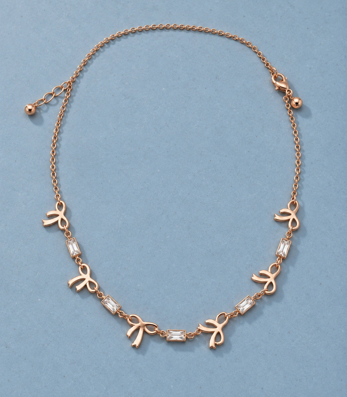 JIMIKI Rose Gold Bow Baguette Crystal Necklace in Rose Gold - editorial flatlay #color_rose-gold