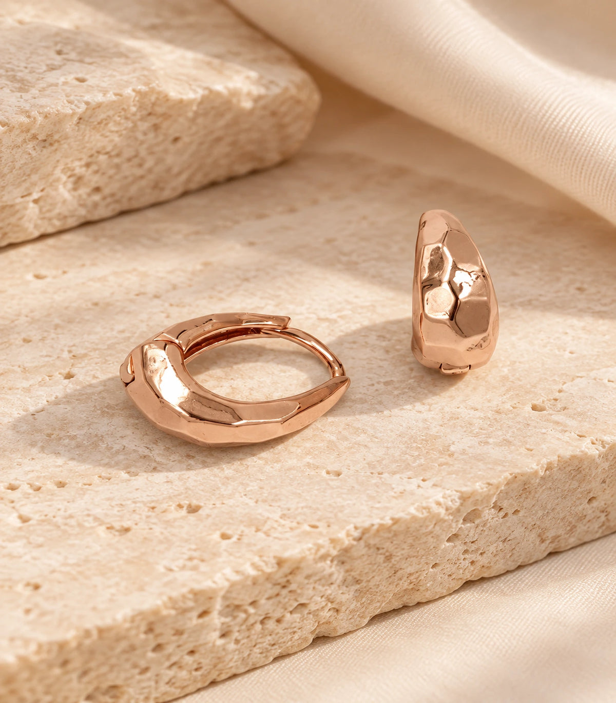 JIMIKI rose gold faceted huggie hoop earrings, pair styled on textured stone #color_rose-gold-tone