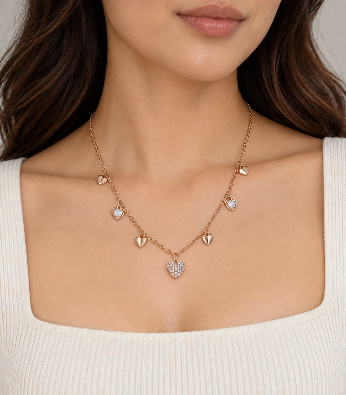 JIMIKI rose gold-tone seven heart charm necklace, necklace worn over an ivory top with the model's eyes outside the frame #color_rose-gold