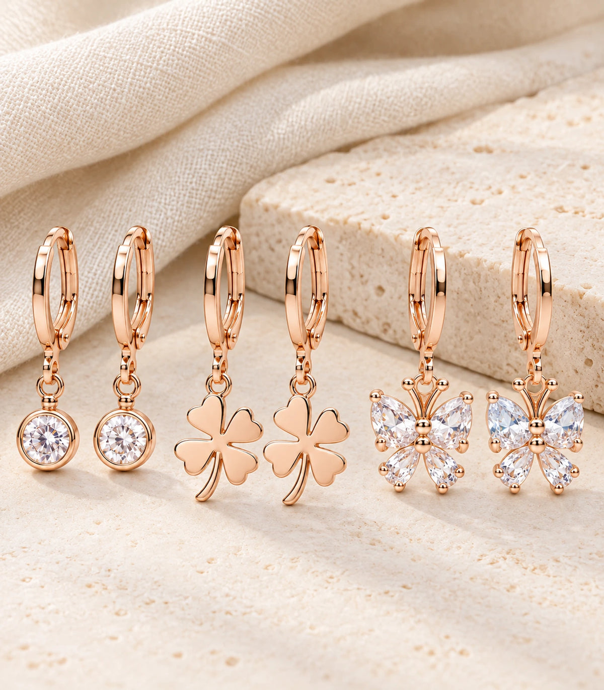 All six rose gold tone crystal, clover and butterfly hoop earrings arranged on ivory linen and pale stone