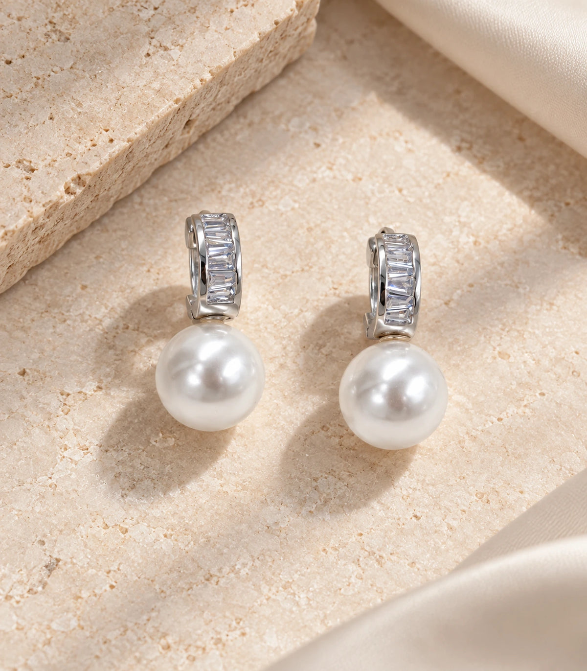 Silver baguette huggie earrings with white pearl-style drops styled as a pair on textured stone