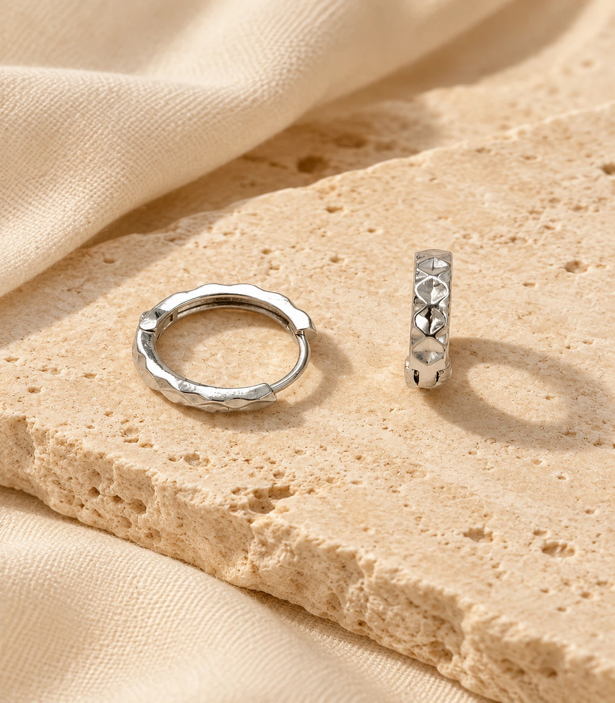 JIMIKI silver faceted diamond pattern huggie earrings, pair styled on textured stone #color_silver