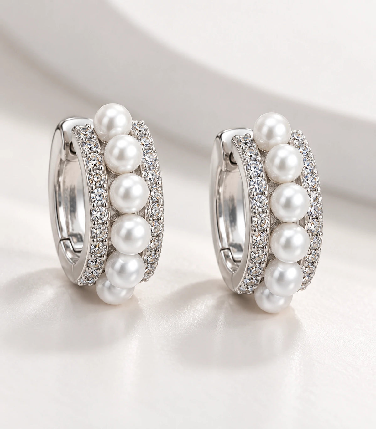 Close view of the matching silver-tone pearl and pavé huggie earring pair on fabric