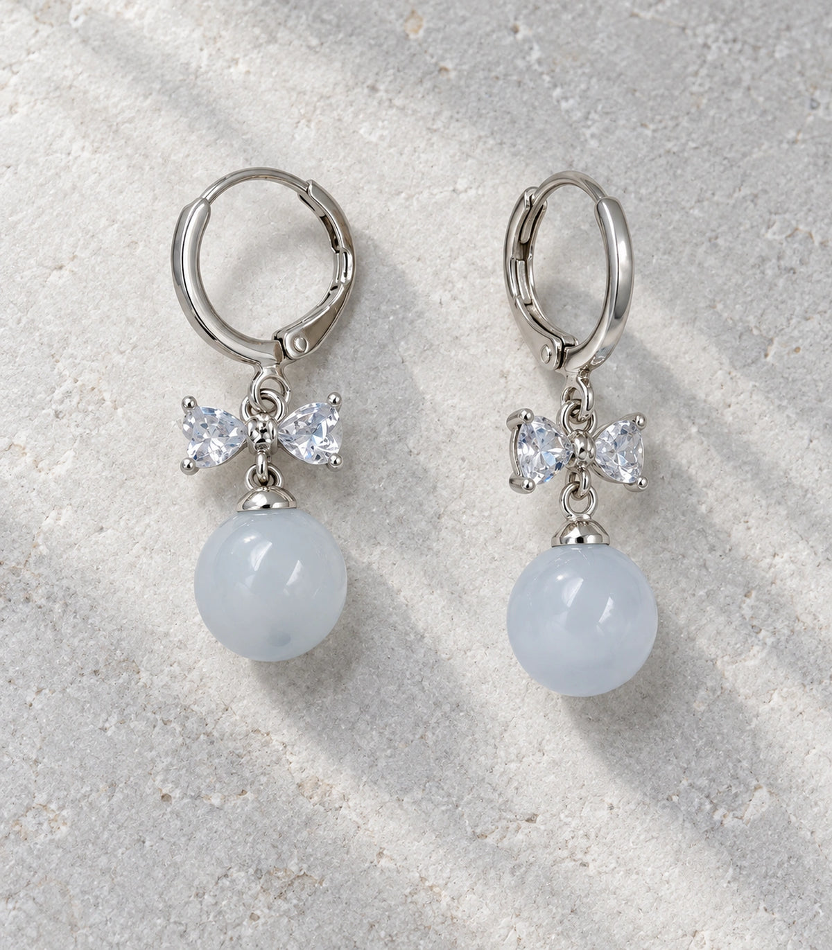 JIMIKI Silver-Tone Clear Crystal Bow Pale Blue Bead-Drop Huggie Earrings in Silver / Clear / Pale Blue, editorial flat lay