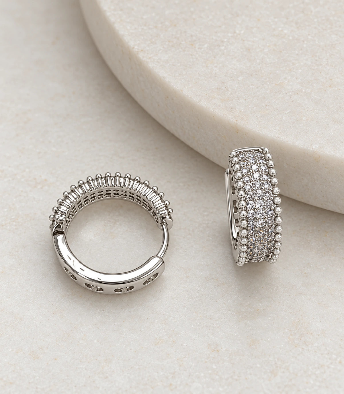 JIMIKI Silver Crystal Bead Border Huggie Hoop Earrings, matching pair on a textured stone surface #color_silver