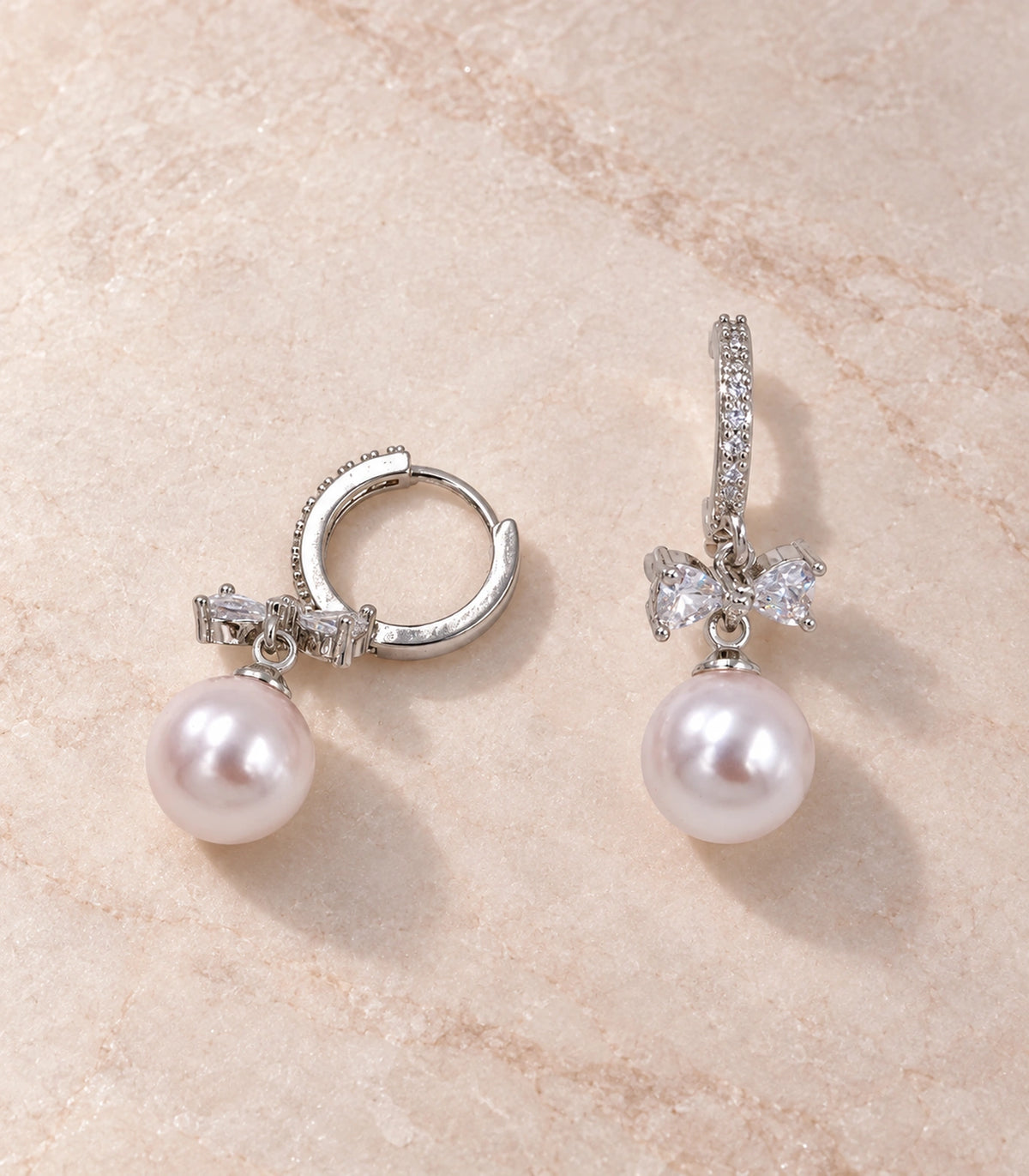 JIMIKI Silver-Tone Crystal Bow Pearl-Drop Huggie Earrings in Silver / Clear / White, editorial flat lay