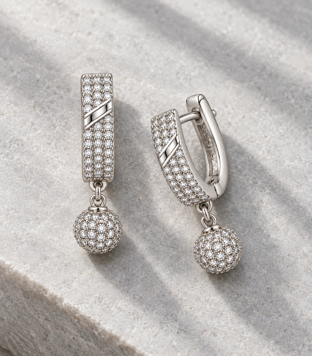 JIMIKI Silver-Tone Diagonal Pavé Ball Drop Huggie Earrings — pair in a styled flat lay #color_silver-tone / clear