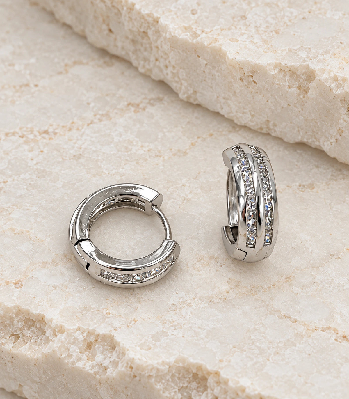 JIMIKI Silver Double Channel Crystal Huggie Hoop Earrings, matching pair on a textured stone surface #color_silver