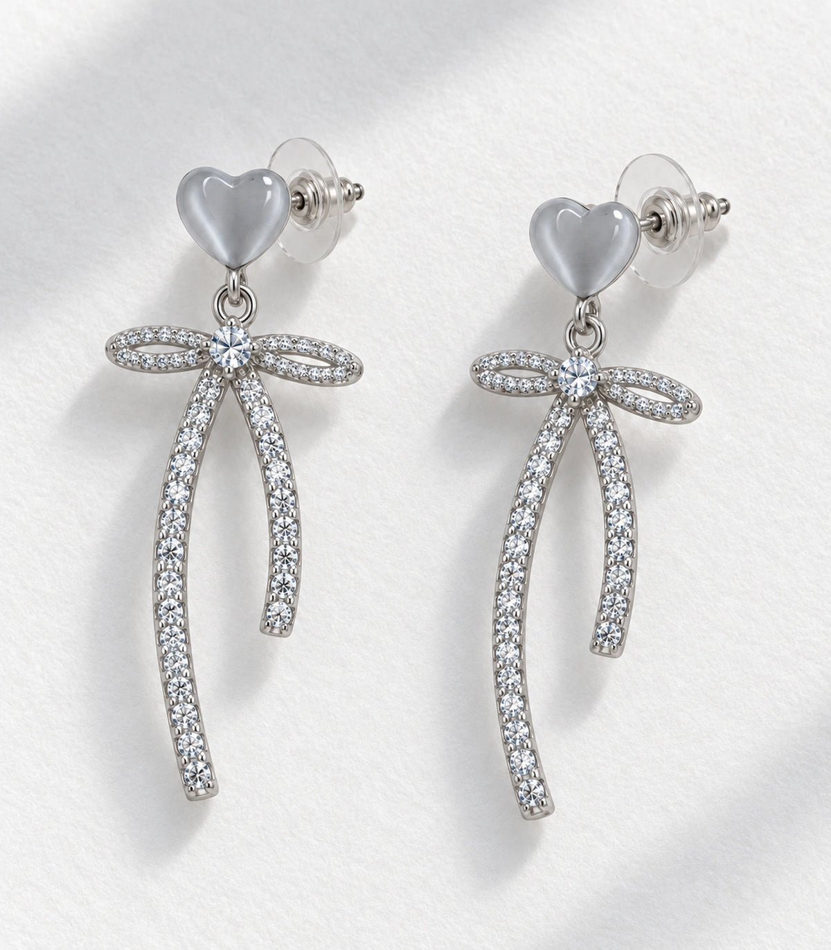 JIMIKI Heart Crystal Ribbon Bow Drop Earring Pair in Silver-tone / Grey, editorial flat lay #color_silver-tone / grey