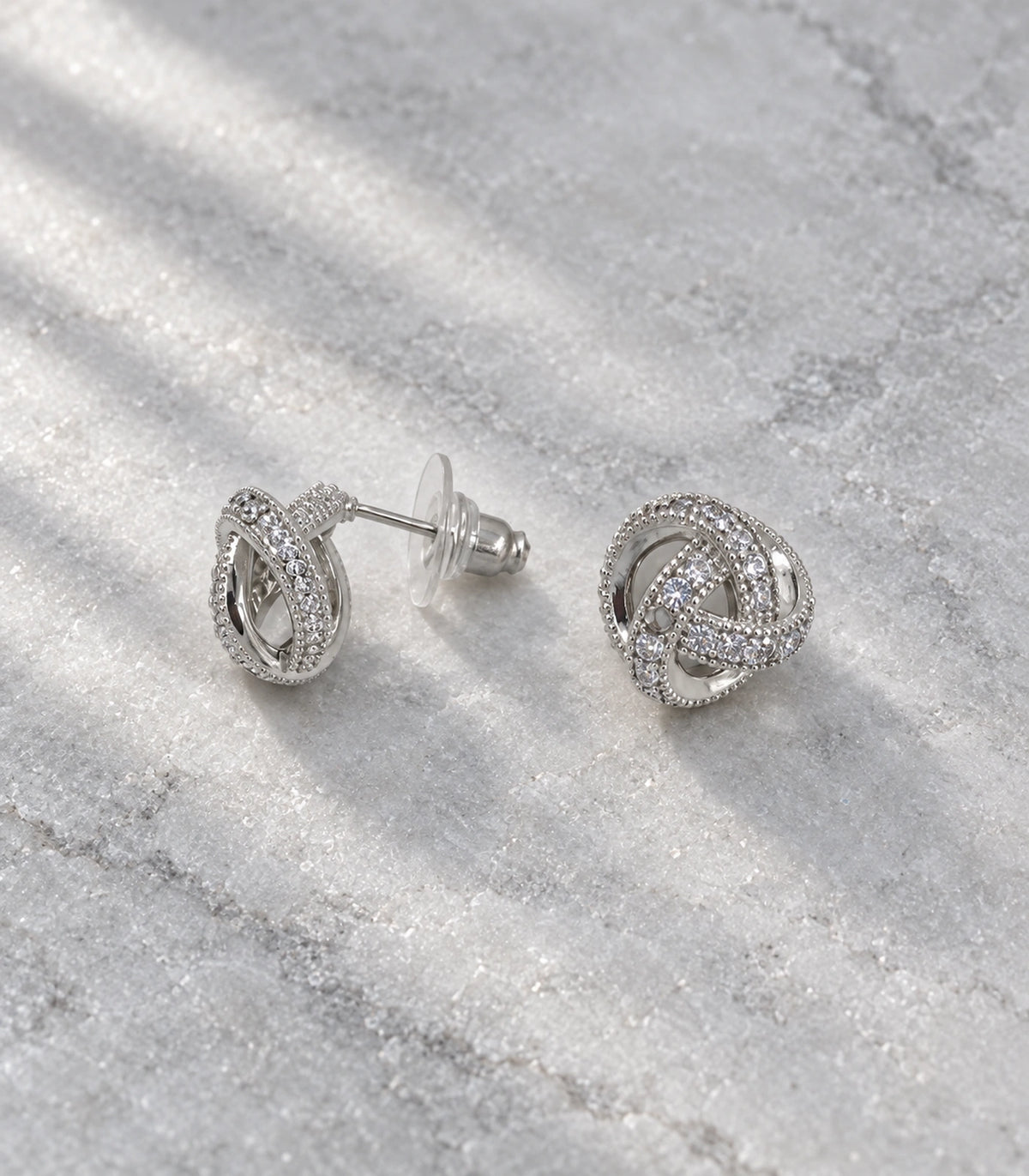 JIMIKI Silver-Tone Pavé Interwoven Knot Stud Earrings in Silver / Clear, editorial flat lay