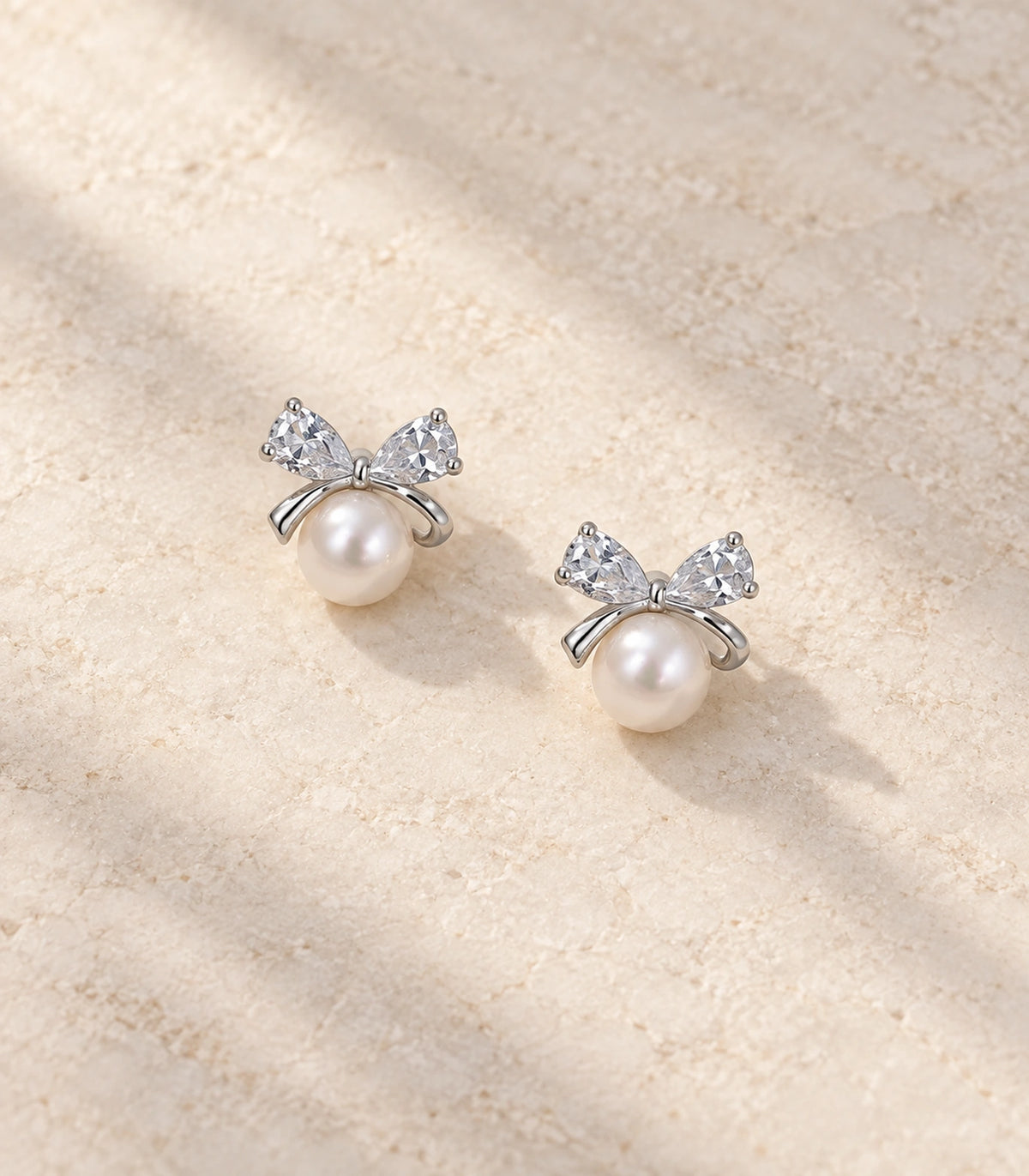 JIMIKI Silver-Tone Pearl Crystal Bow Stud Earrings in Silver / White, editorial flat lay