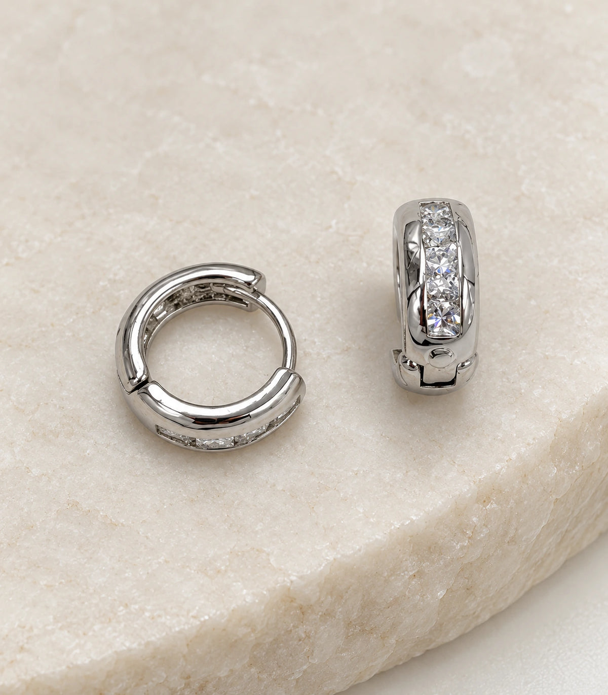 JIMIKI Silver Rectangular Crystal Channel Huggie Hoop Earrings, matching pair on a textured stone surface #color_silver