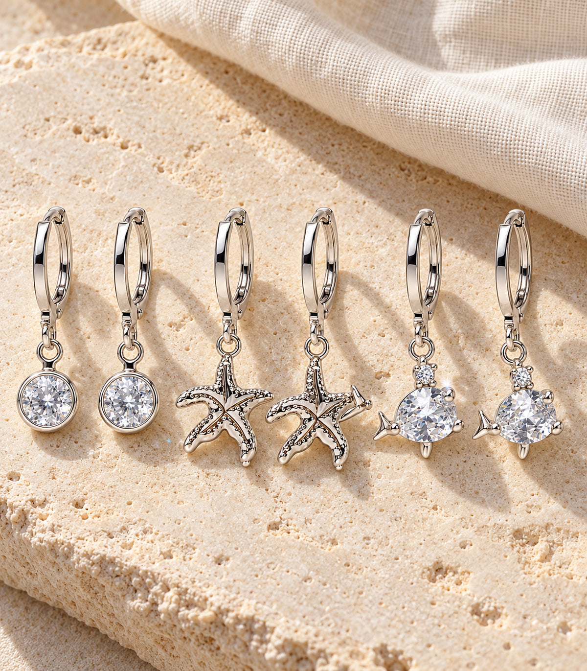 All three matching silver-tone hoop earring pairs with round crystal, starfish and fish charms on stone and linen.