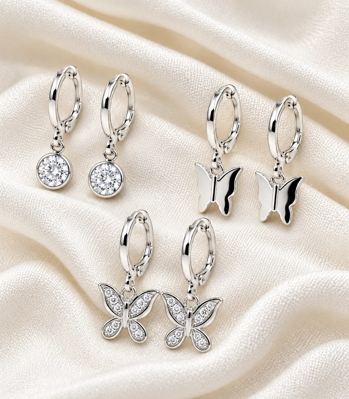 Three pairs of silver-tone crystal and butterfly hoop earrings arranged on soft ivory fabric