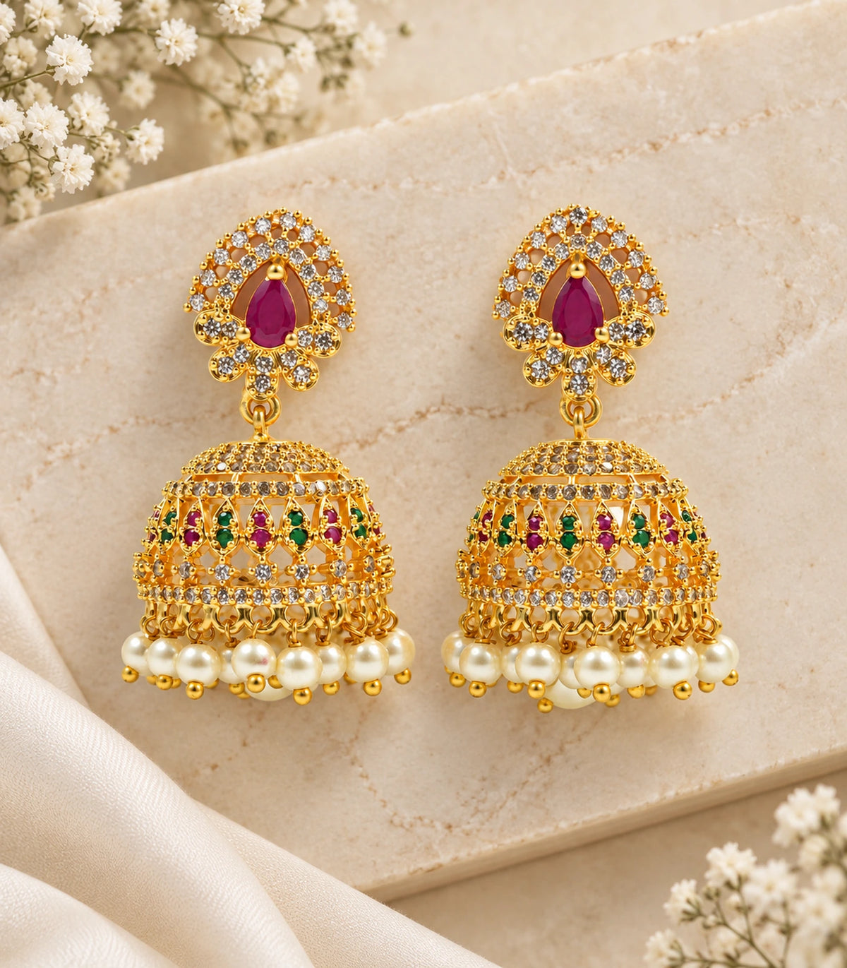 Complete pair of gold-tone magenta and green lattice pearl-look jhumka earrings styled on cream stone
