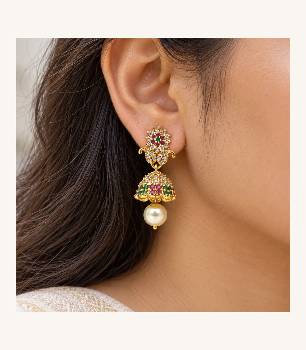 Closeup of one magenta and green gold-tone earring worn at the ear, showing floral stones, scrolls, dome and pearl-like drop
