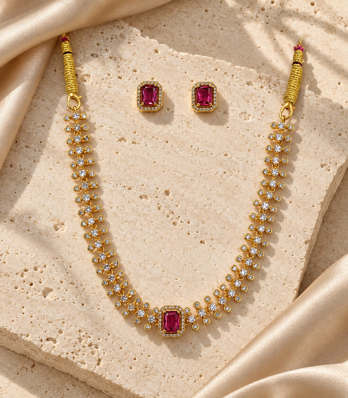 Gold-tone magenta crystal necklace and two matching stud earrings arranged on stone and fabric