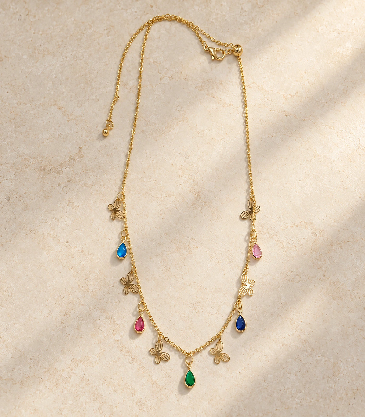 JIMIKI Gold Multicolor Teardrop Butterfly Charm Necklace - complete necklace on a pale textured surface #color_gold