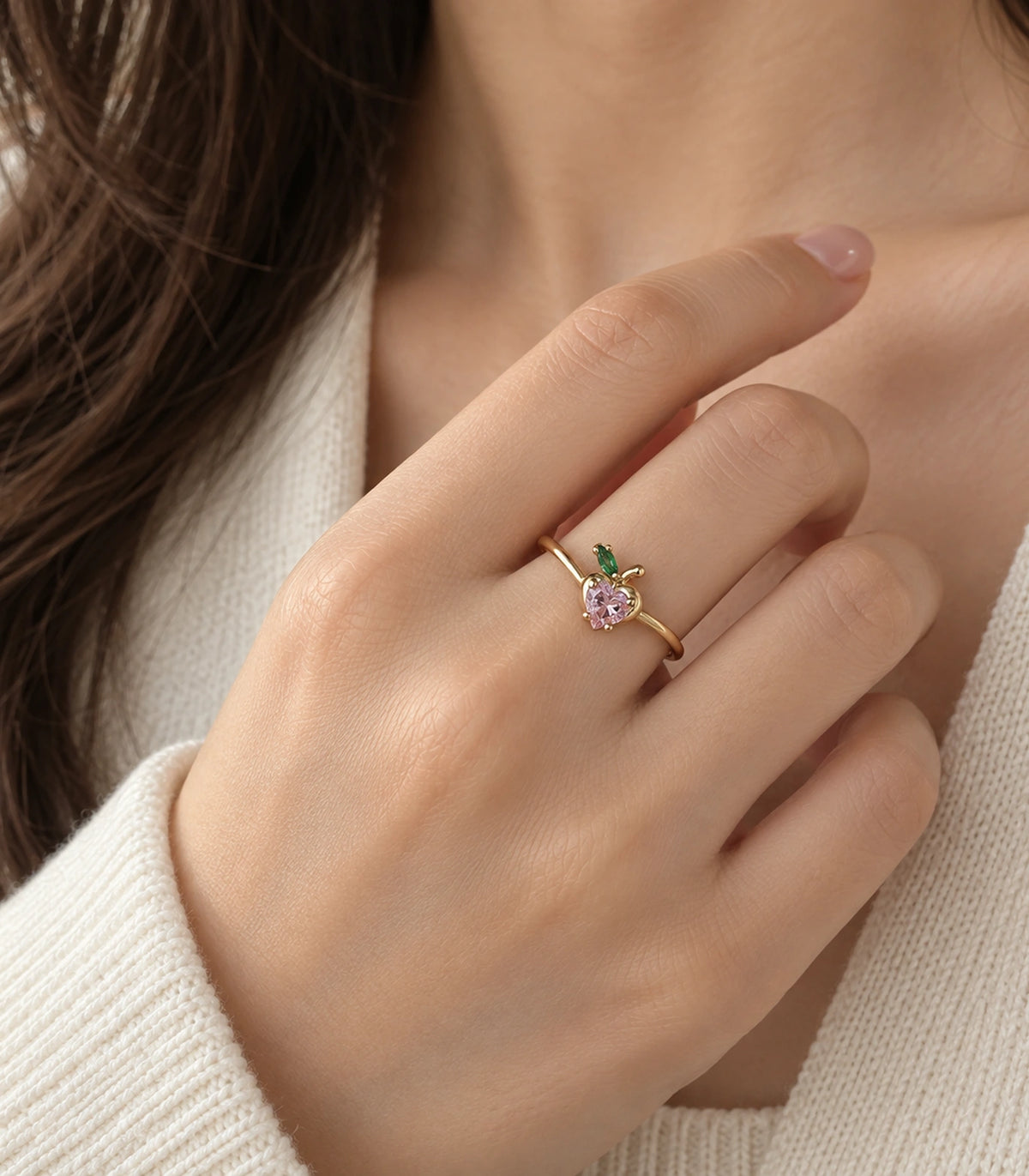 Gold pink heart and green leaf ring worn in a hand-at-neck pose with cream knit styling
