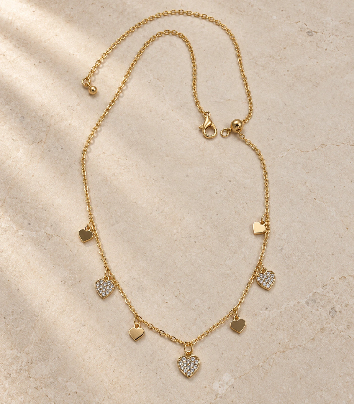 JIMIKI adjustable gold-tone seven heart charm necklace, full necklace on textured stone showing the clasp-side adjustment bead #color_gold