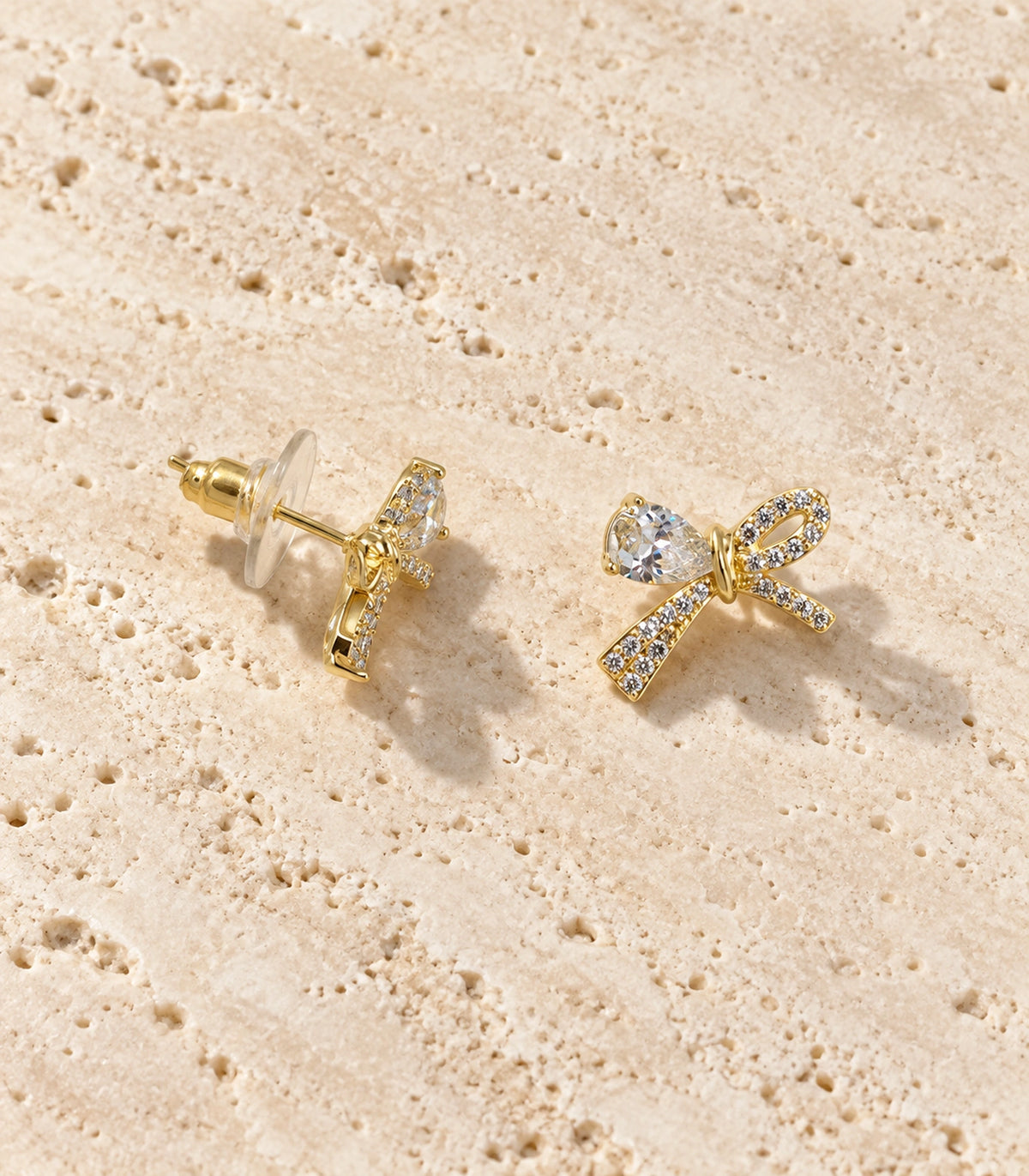 JIMIKI Gold-Tone Asymmetric Crystal Bow Stud Earrings in Gold / Clear, editorial flat lay
