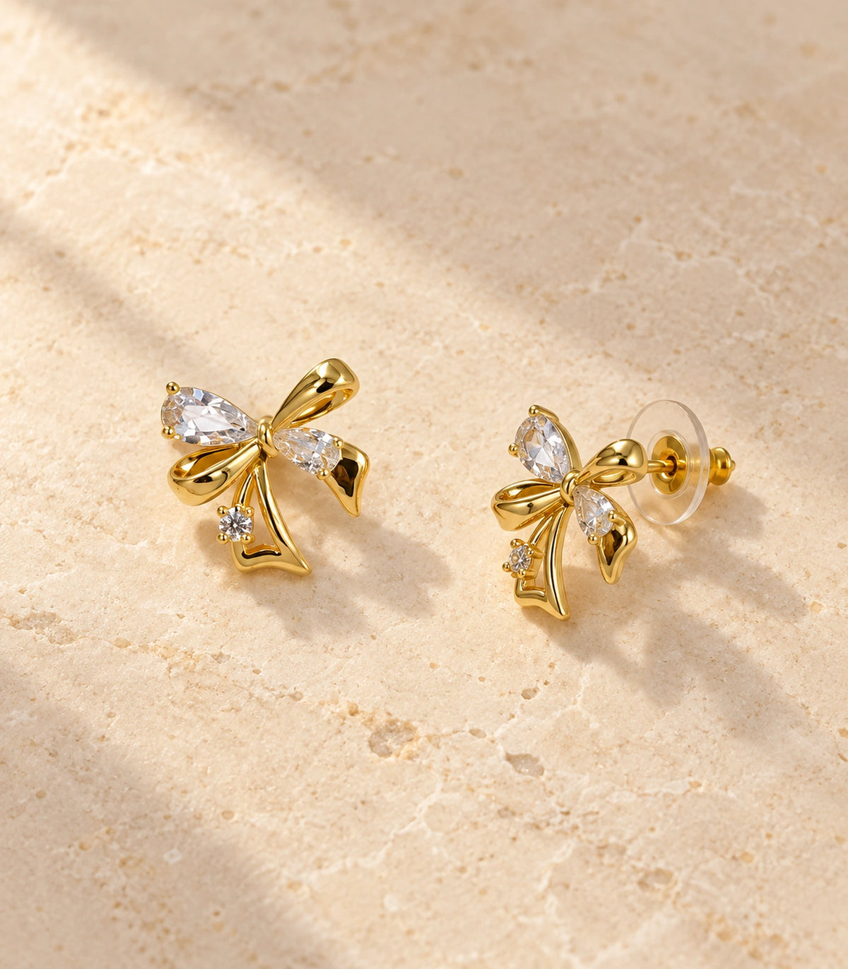 JIMIKI Asymmetric Crystal Bow Stud Earring Pair in Gold / Clear, editorial flat lay #color_gold / clear