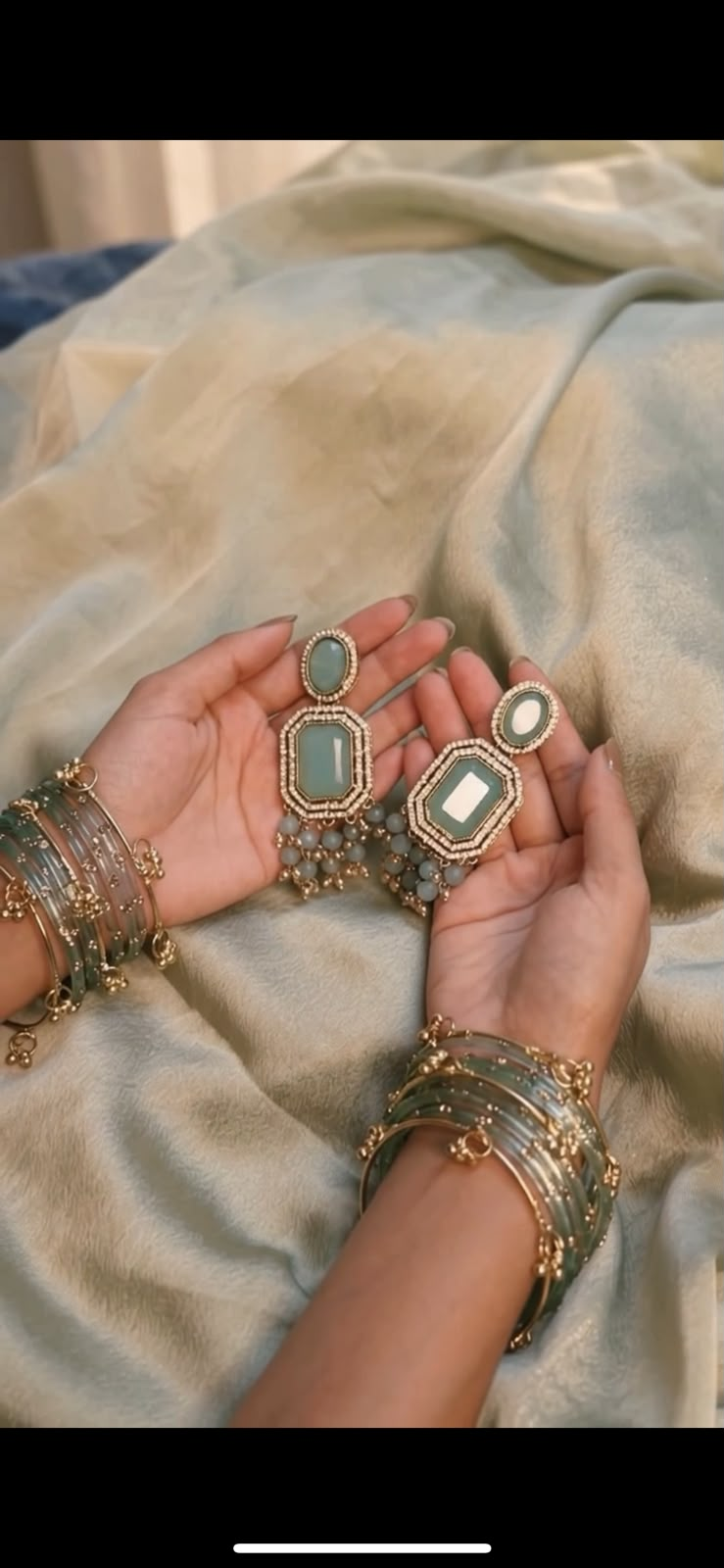 Hands holding sage-green statement earrings with matching bangles on soft ivory fabric