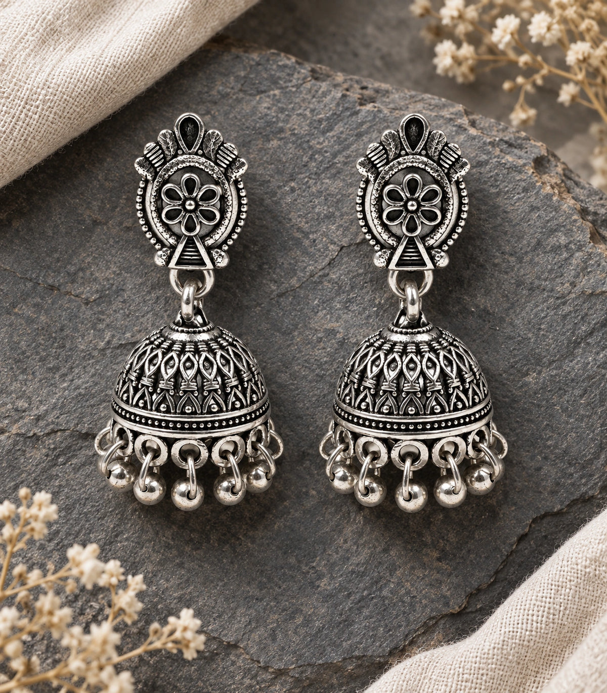 JIMIKI Oxidized Silver Oval Floral Jhumka Earrings - Matching pair on grey stone beside cream fabric #color_oxidized-silver