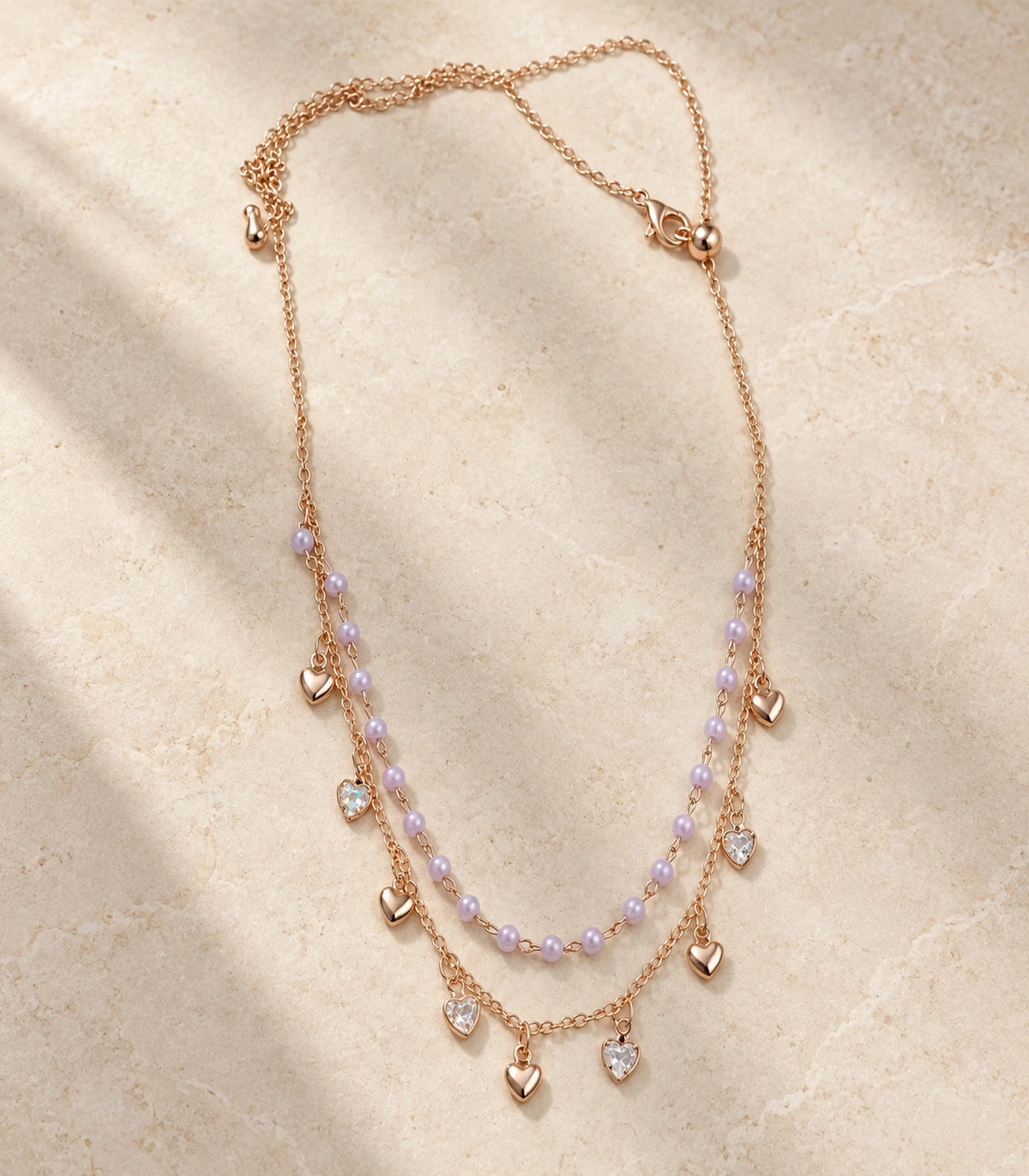 JIMIKI Rose Gold Lavender Bead Heart Charm Layered Necklace - complete necklace on a pale textured surface #color_rose-gold