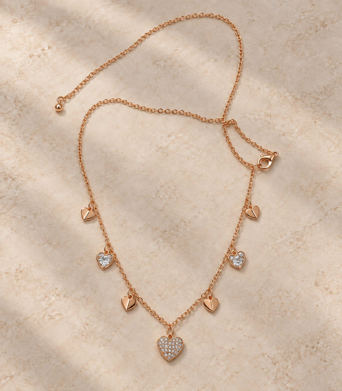 JIMIKI rose gold-tone seven heart charm necklace, necklace arranged on a neutral surface showing the seven heart charms #color_rose-gold
