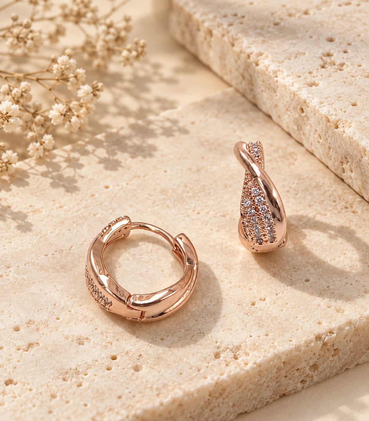 JIMIKI Rose Gold Tone crossover pave huggie hoop earrings, pair arranged on textured stone #color_rose-gold-tone