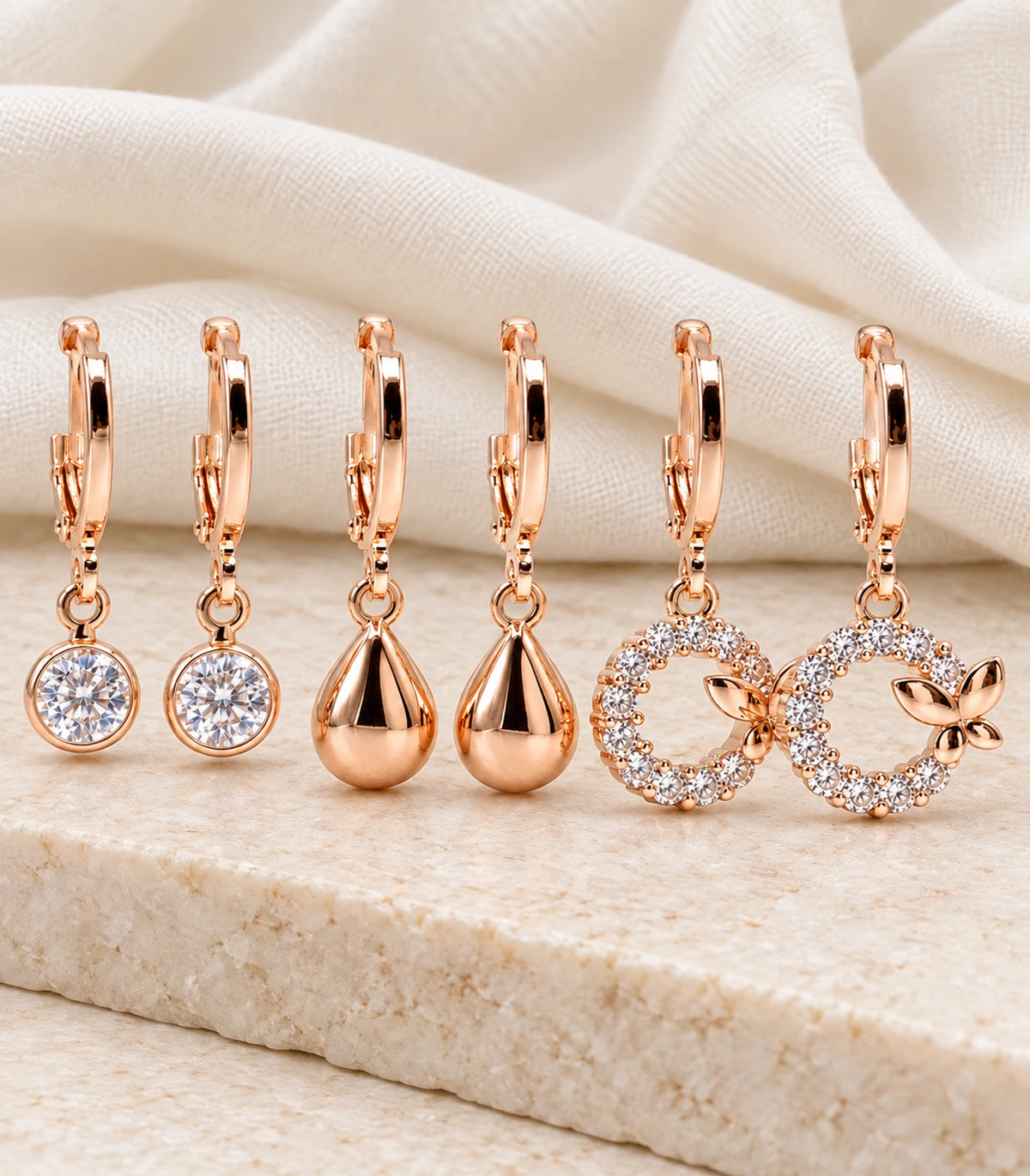 Complete three-pair rose gold tone crystal, teardrop and four-leaf wreath hoop earring set on pale fabric