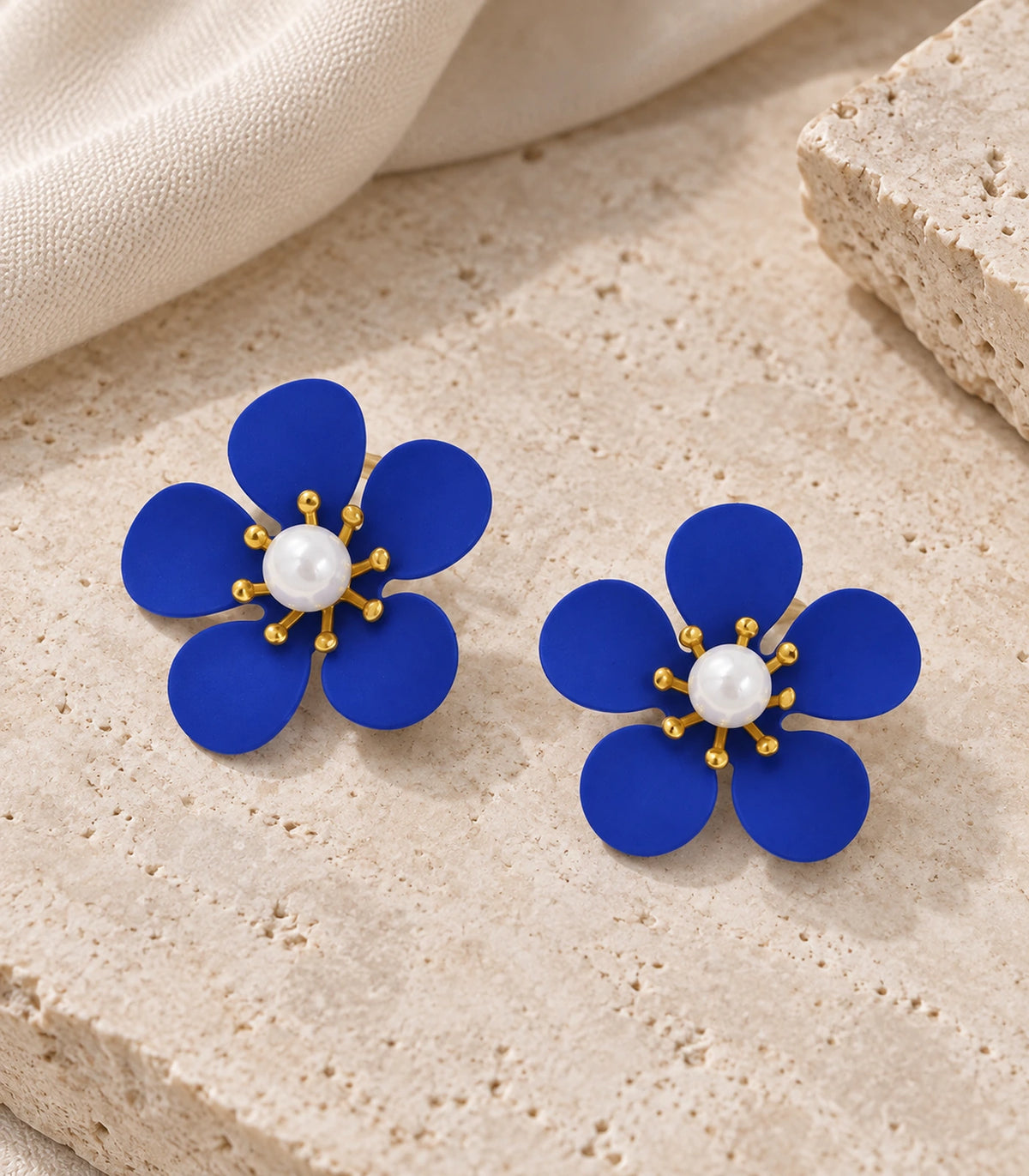 JIMIKI royal blue five-petal flower stud earrings with pearl-effect centre, matching flower stud pair in an editorial flat lay #color_royal-blue