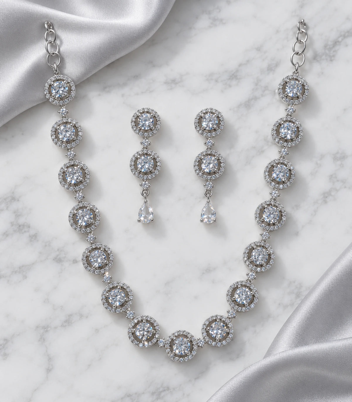 Silver-tone clear round halo necklace and matching pair of pear-drop earrings arranged on marble with fabric